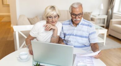Happy middle aged husband and wife sitting at table with laptop and paper bills, calculating domestic incomes together at home.